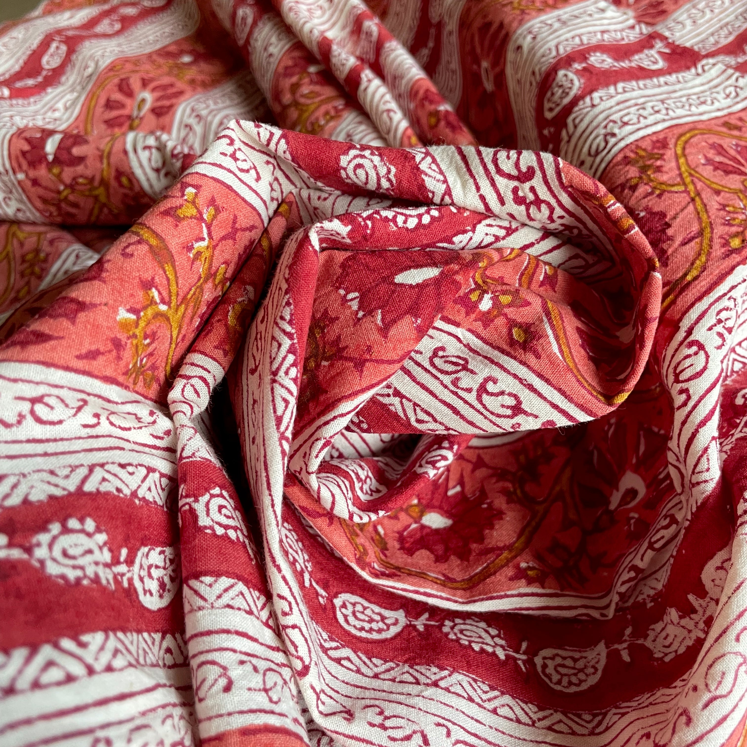 A close-up of draped fabric with a red and white block-printed floral and geometric pattern, featuring warm orange-yellow accents.