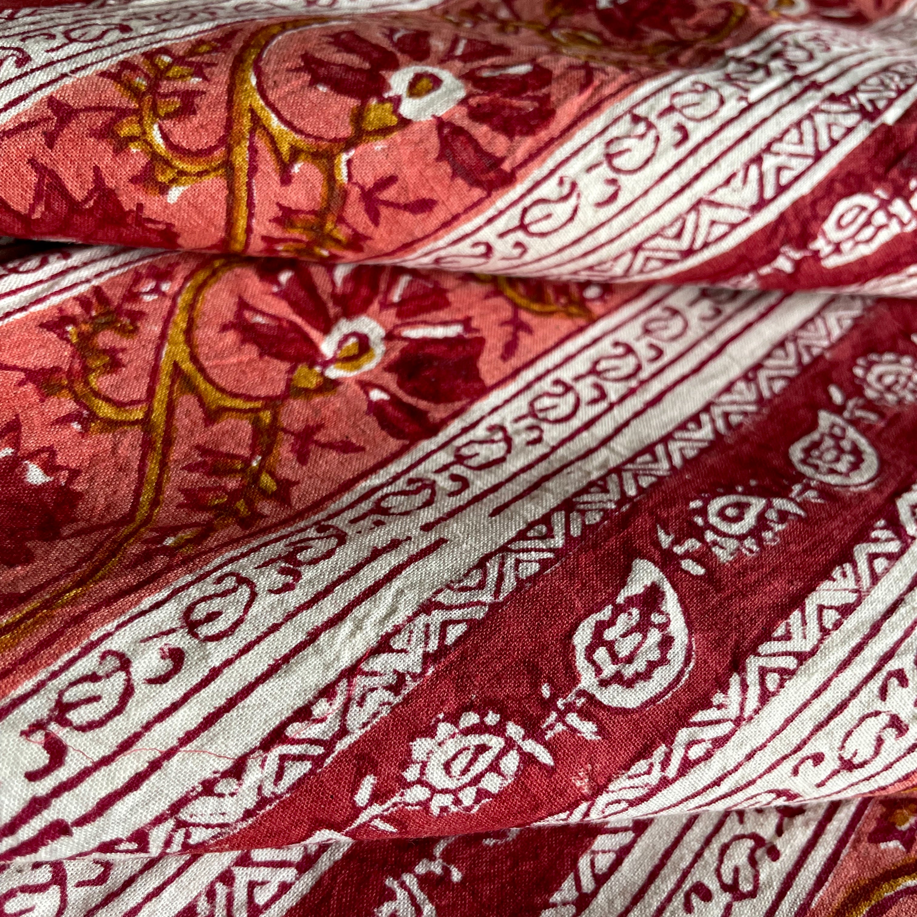 A close-up of folded patterned fabric with a coral pink base, deep red floral and border motifs, and mustard-gold decorative accents.