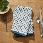 Stack of blue and white patterned napkins on a wooden table with a fork, knife, and herb bowl.